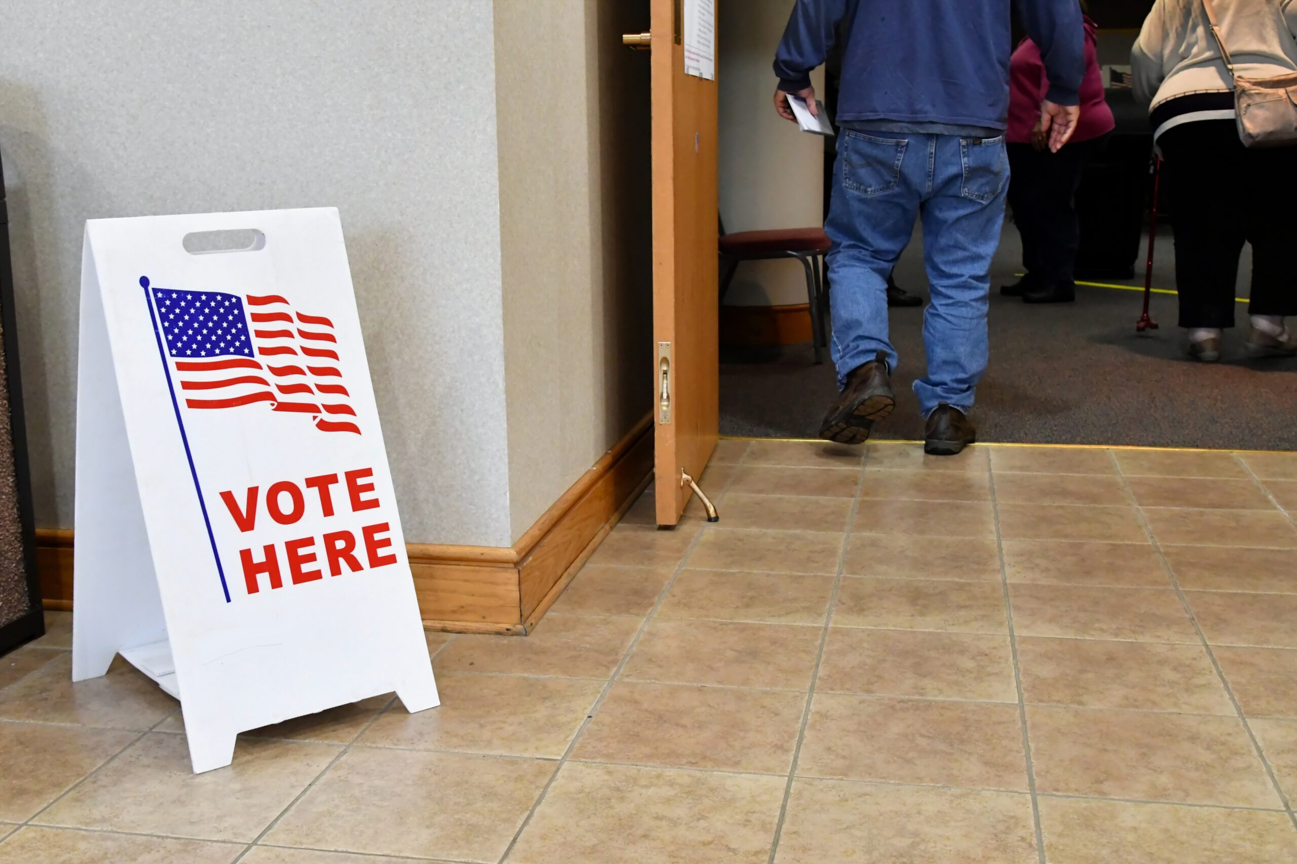 "Vote Here" sign inside polling place; people queued to vote