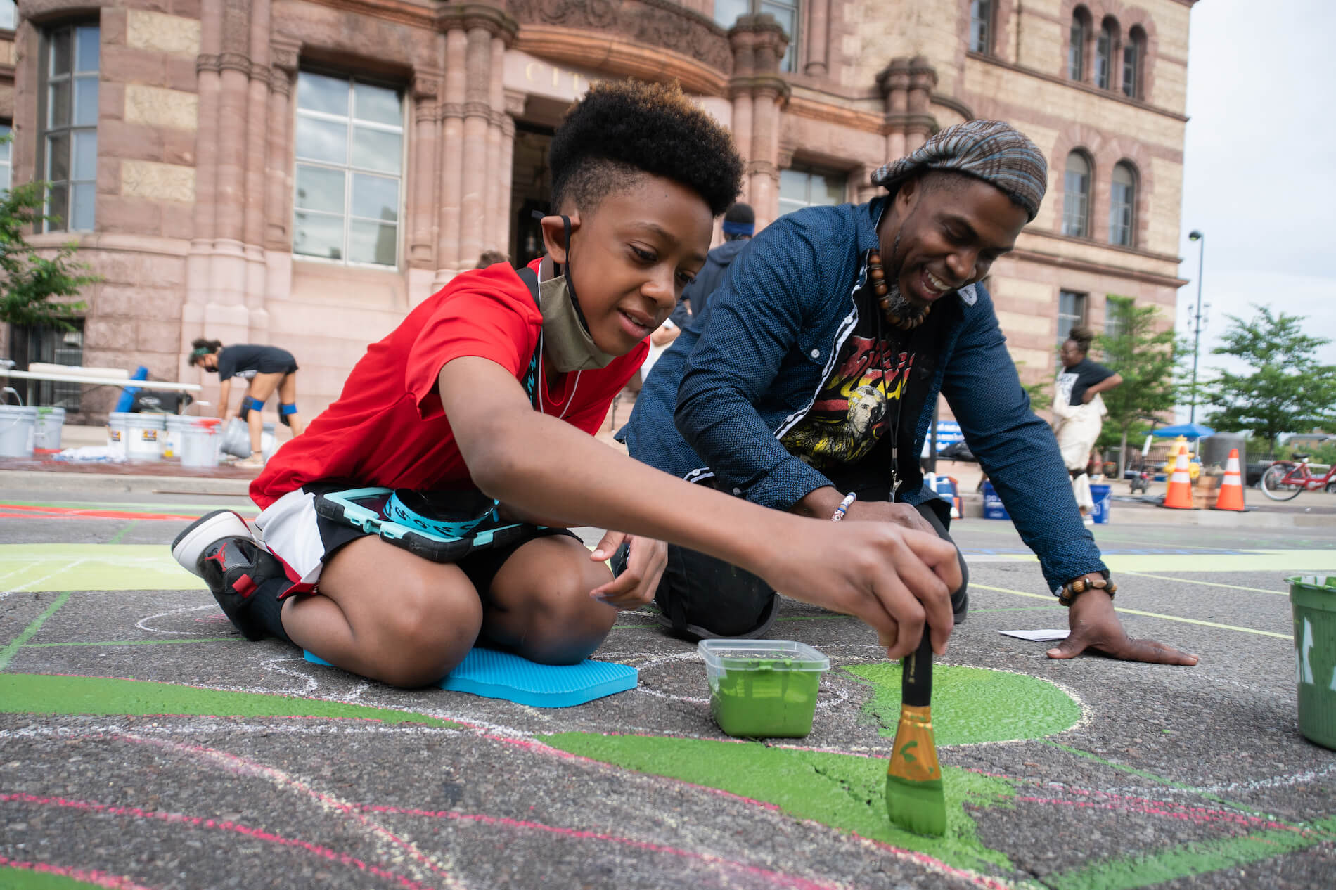 Black father and son painting mural on ground.