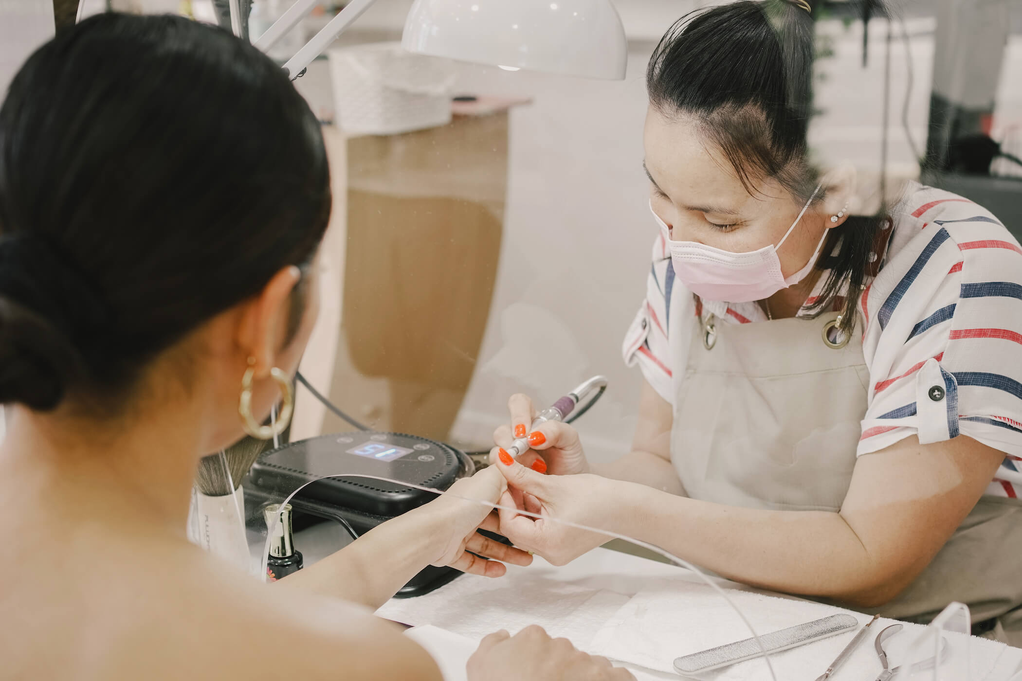 Asian manicure therapist filing customer nails in nail salon with protective screen