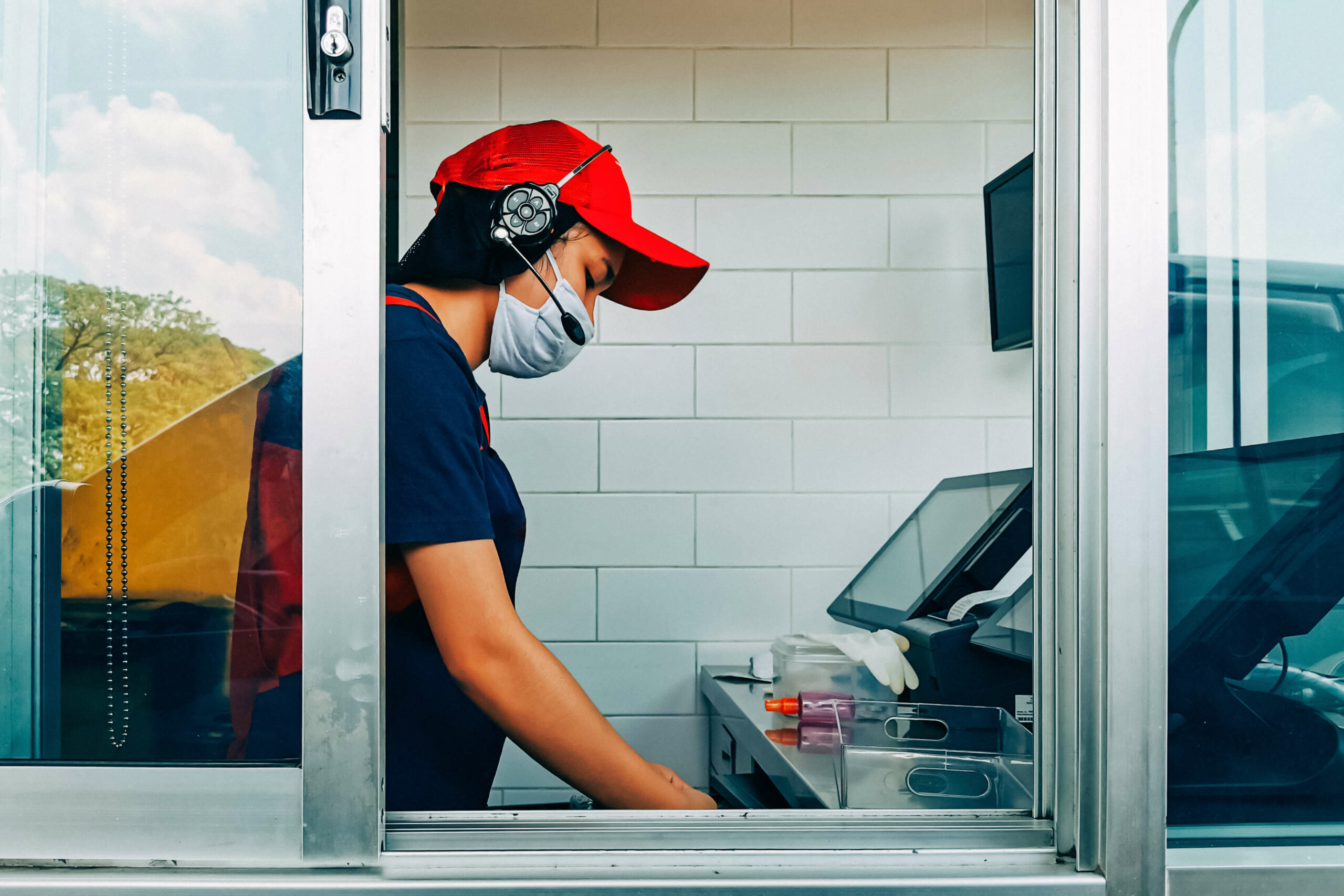Fast food worker wearing masks opens cash register at drive-through window Gumpanat / Shutterstock.com